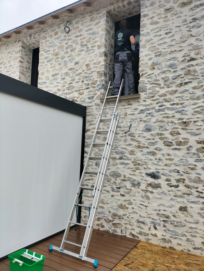 Employé Familles & Services en intervention sur une maison en pierre — échelle, tenue logo, bac vert